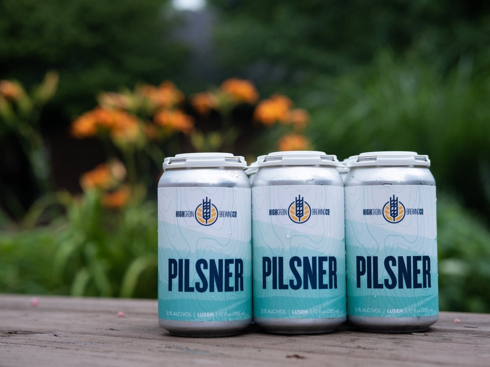 A six-pack of HighGrain Lusen Pilsner cans on a wooden table in front of orange lilies