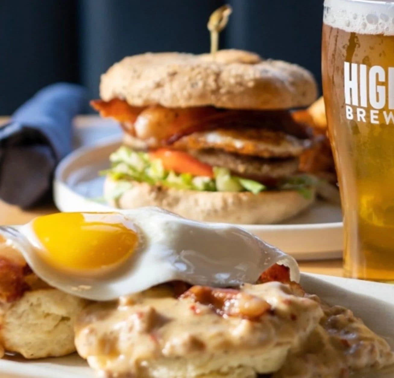 Close-up of a breakfast dish with a fried egg on top of biscuits and gravy in the foreground, and a large burger with bacon, lettuce, tomato, and a sandwich bun in the background. A glass of beer is partially visible on the right with the word 'HIGH' on it.