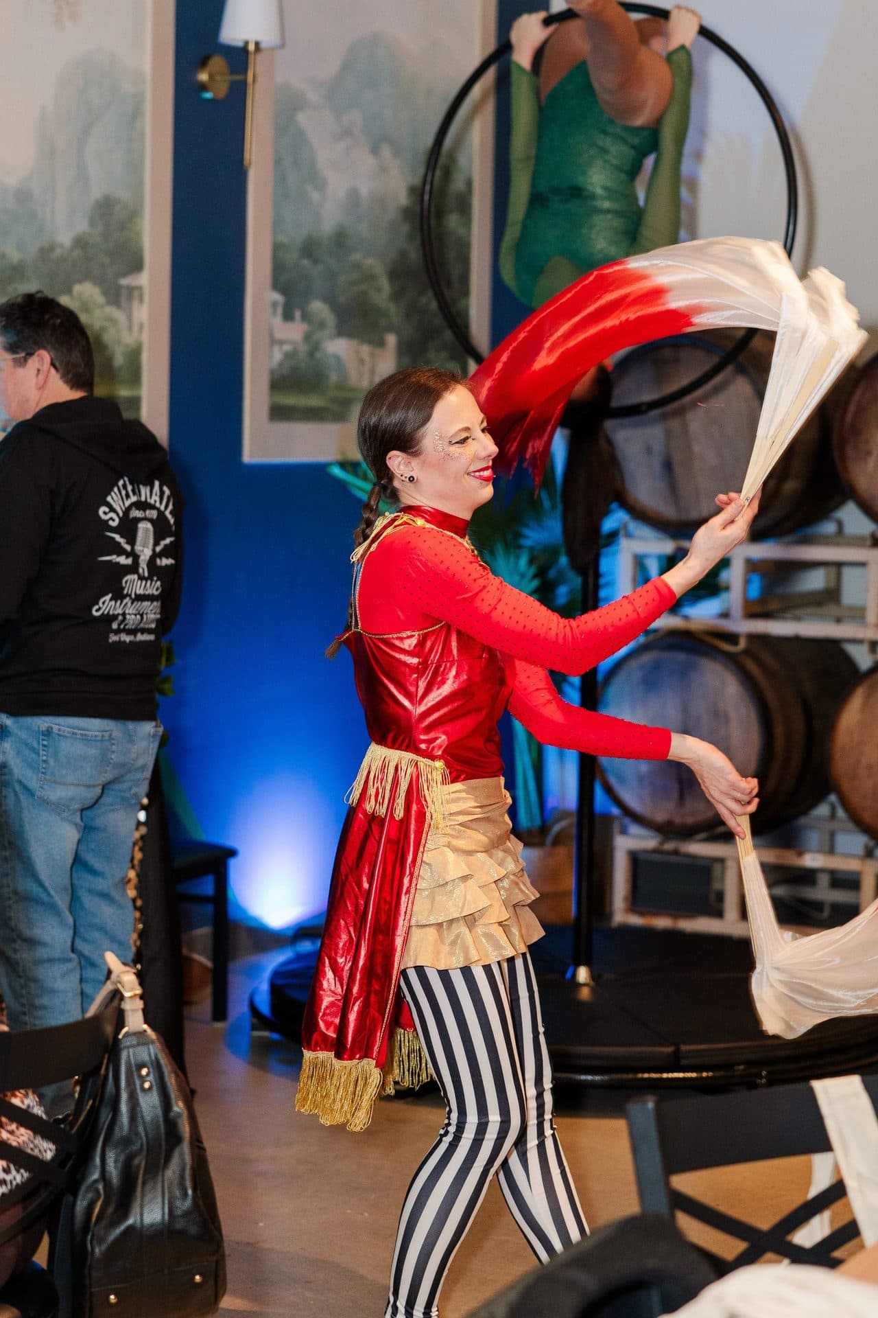A woman in a colorful costume performing a circus act with a fan of fabric, smiling with lipstick, with people and decorations in the background.