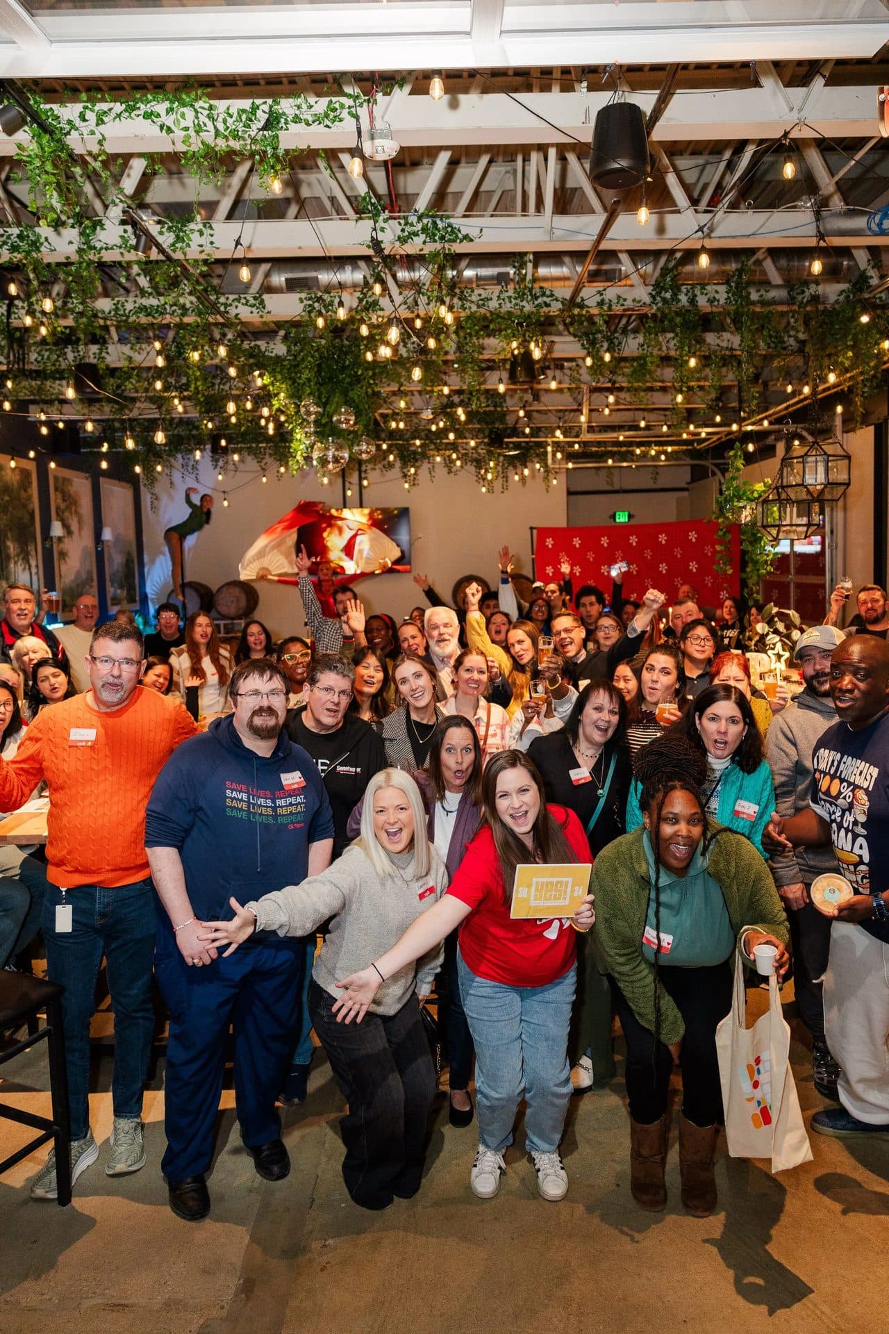 A large group of people gathered indoors at a celebration or event, with some raising their hands and smiling. The ceiling is decorated with string lights and greenery. There are paintings on the walls and a television screen in the background. The crowd appears happy and engaged.