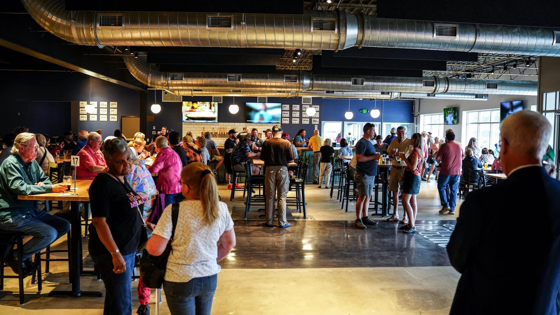 Crowded bar or restaurant interior with people socializing, sitting at high tables and bar counters, large air ducts overhead, multiple TV screens on dark-colored walls, and large windows letting in natural light.